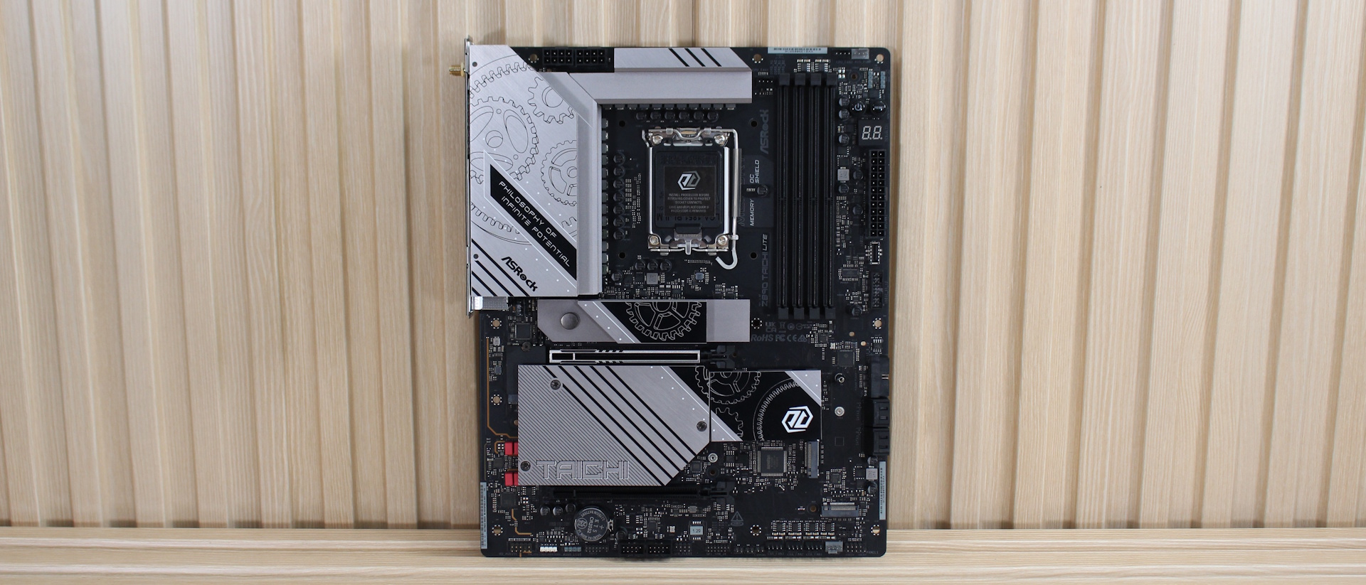 ASRock Z890 Taichi Lite прислонена к решетчатой стене перед проведением тестов производительности. Хотя эта "более низкая" модель Taichi не имеет встроенной RGB-подсветки, ее серебристо-черный вид с мотивом шестерен, охватывающим несколько радиаторов, помогает ей выделяться среди конкурентов.
